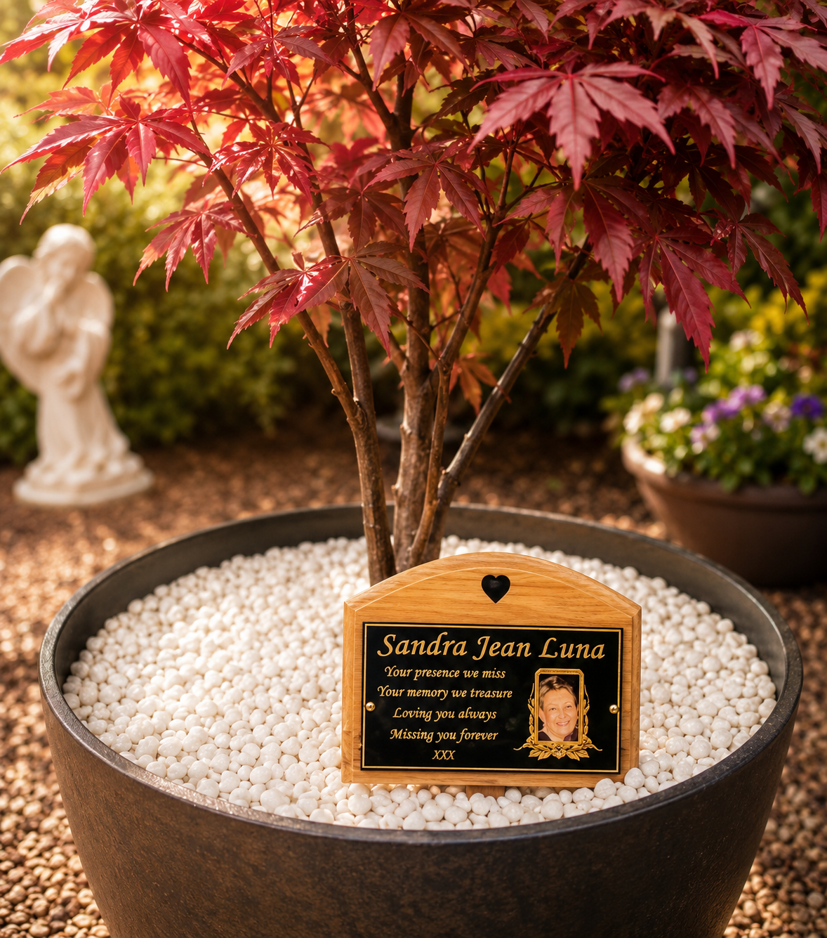 Personalized wooden memorial plaque for a pet, displayed in a black planter filled with white pebbles next to a red-leafed tree in an outdoor garden setting.