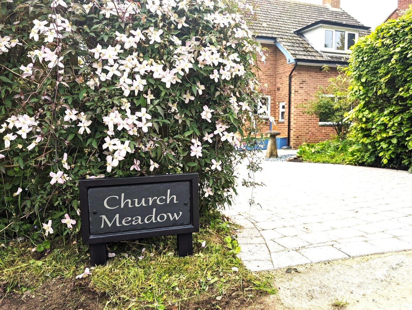 A black rectangular sign with white text reading 'Church Meadow' placed in a garden beside a large flowering bush with pale pink and white blossoms, with a brick house and paved driveway in the background.