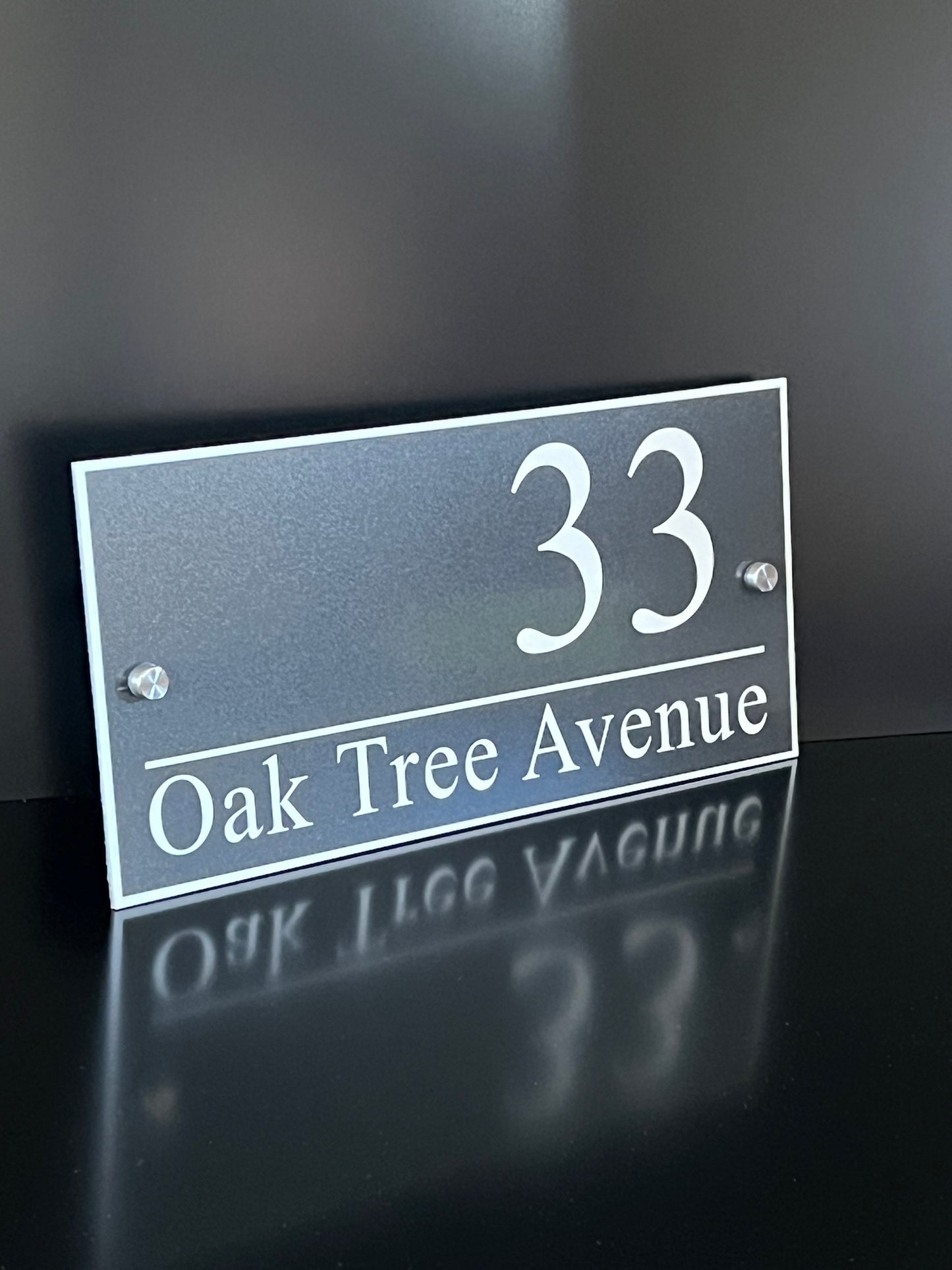 A rectangular metallic house number sign reading '33 Oak Tree Avenue' in white lettering, mounted against a dark wall with a reflective surface below.