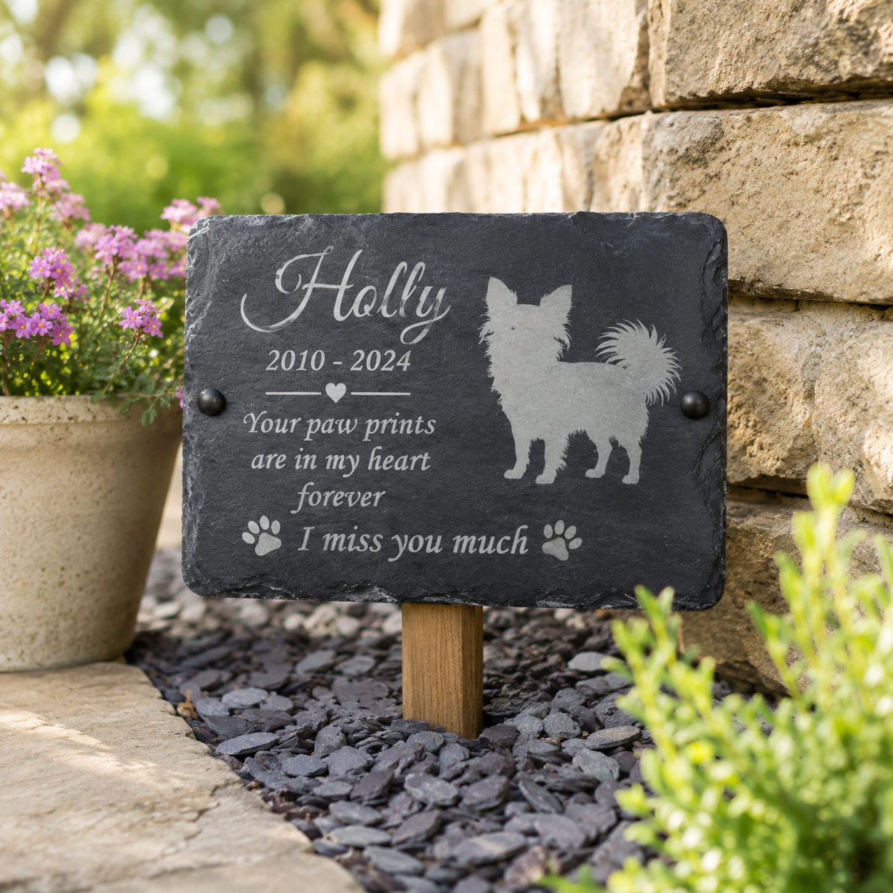 Personalized black slate pet memorial garden stake with engraved text and dog silhouette, mounted on a wooden stake in a garden bed with stone wall and potted flowers in the background.
