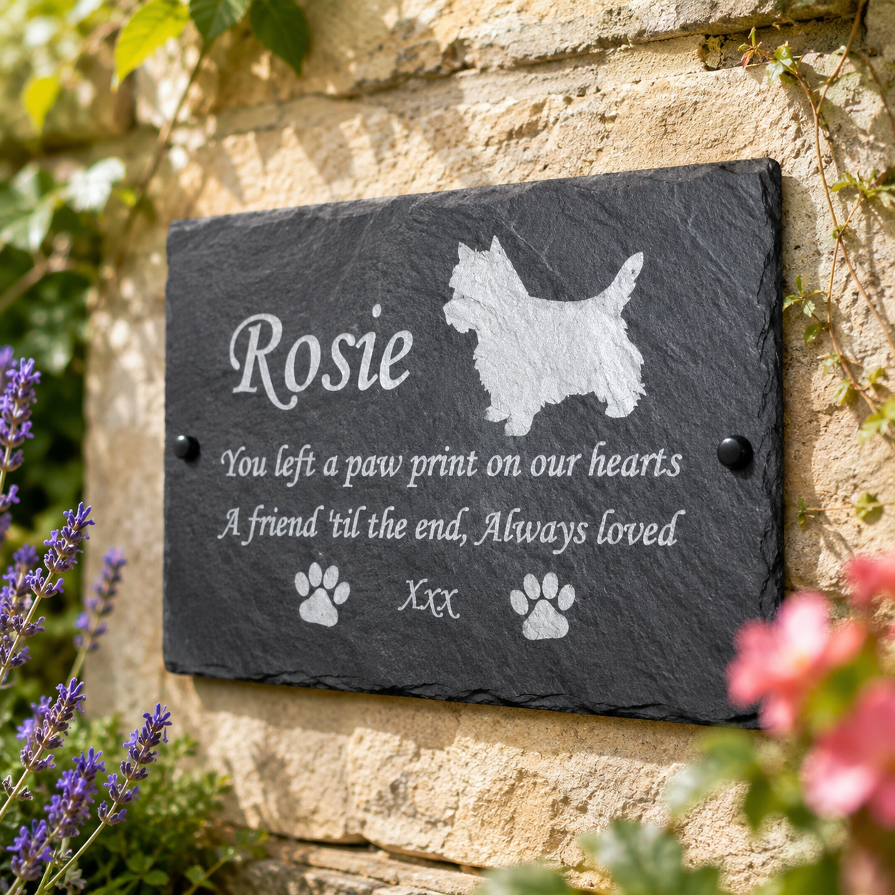 Personalized dark gray slate memorial plaque mounted on a stone wall, engraved with the name Rosie, a West Highland terrier silhouette, paw prints, and a remembrance message, with flowers and greenery in the foreground.