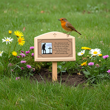 Wooden grave marker with engraved design and text, surrounded by flowers in a garden.