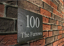 A slate house sign with the number 100 and the text 'The Furrows' engraved on it, mounted on a brick wall.