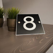 Black plaque with the number '8' on a wooden surface with plants in the background