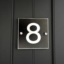 Black square house number plate with the number '8' on a dark wooden background
