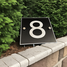 Black square house number '8' sign on a stone ledge with greenery in the background