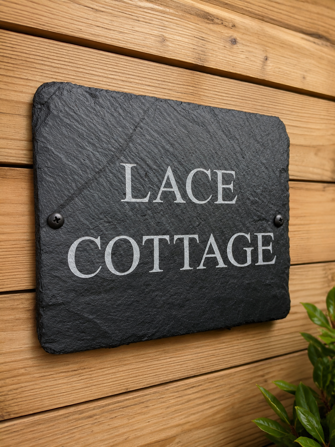 A rectangular dark slate house sign with rounded corners and the words 'LACE COTTAGE' in white serif lettering, mounted on a light wood plank wall with small dark screws at the corners.