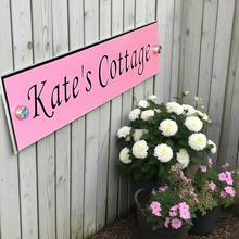 Pink sign with 'Kate's Cottage' text next to potted plants against a wooden fence.