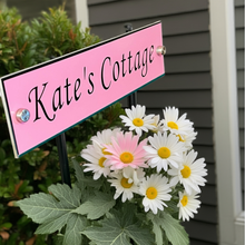 Pink sign with 'Kate's Cottage' text surrounded by flowers in front of a house.
