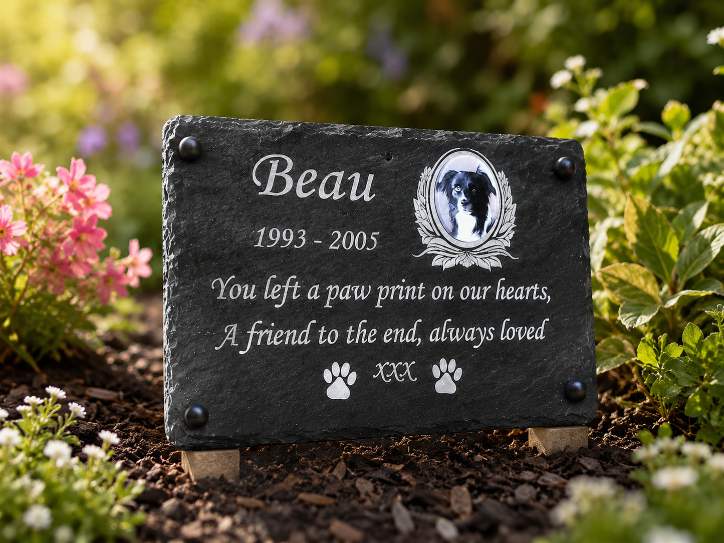 Personalized black slate pet memorial garden marker with engraved white text, a dog portrait, and paw prints, displayed outdoors on a garden bed with flowers and mulch.