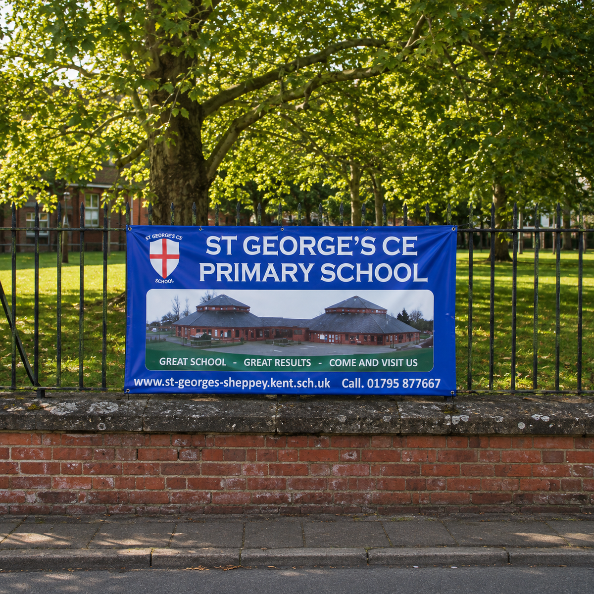 A blue banner for St George's CE Primary School is displayed on a brick wall behind a black metal fence, with a large green tree in the background.