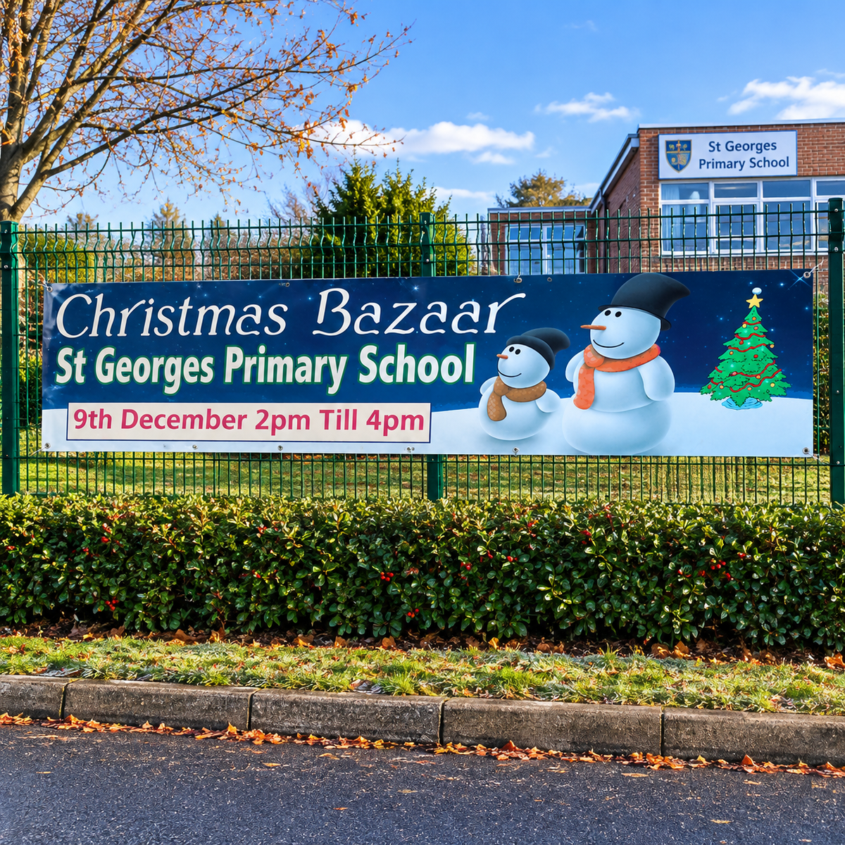 A large outdoor banner for 'Christmas Bazaar St Georges Primary School' displayed on a green fence. The banner features snowman illustrations, a Christmas tree, and event details for December 9th from 2pm to 4pm, with St Georges Primary School building visible in the background under a blue sky.