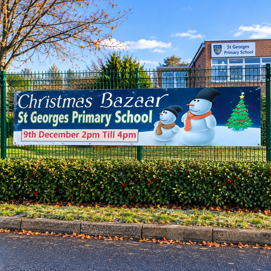 A large outdoor banner for 'Christmas Bazaar St Georges Primary School' displayed on a green fence. The banner features snowman illustrations, a Christmas tree, and event details for December 9th from 2pm to 4pm, with St Georges Primary School building visible in the background under a blue sky.