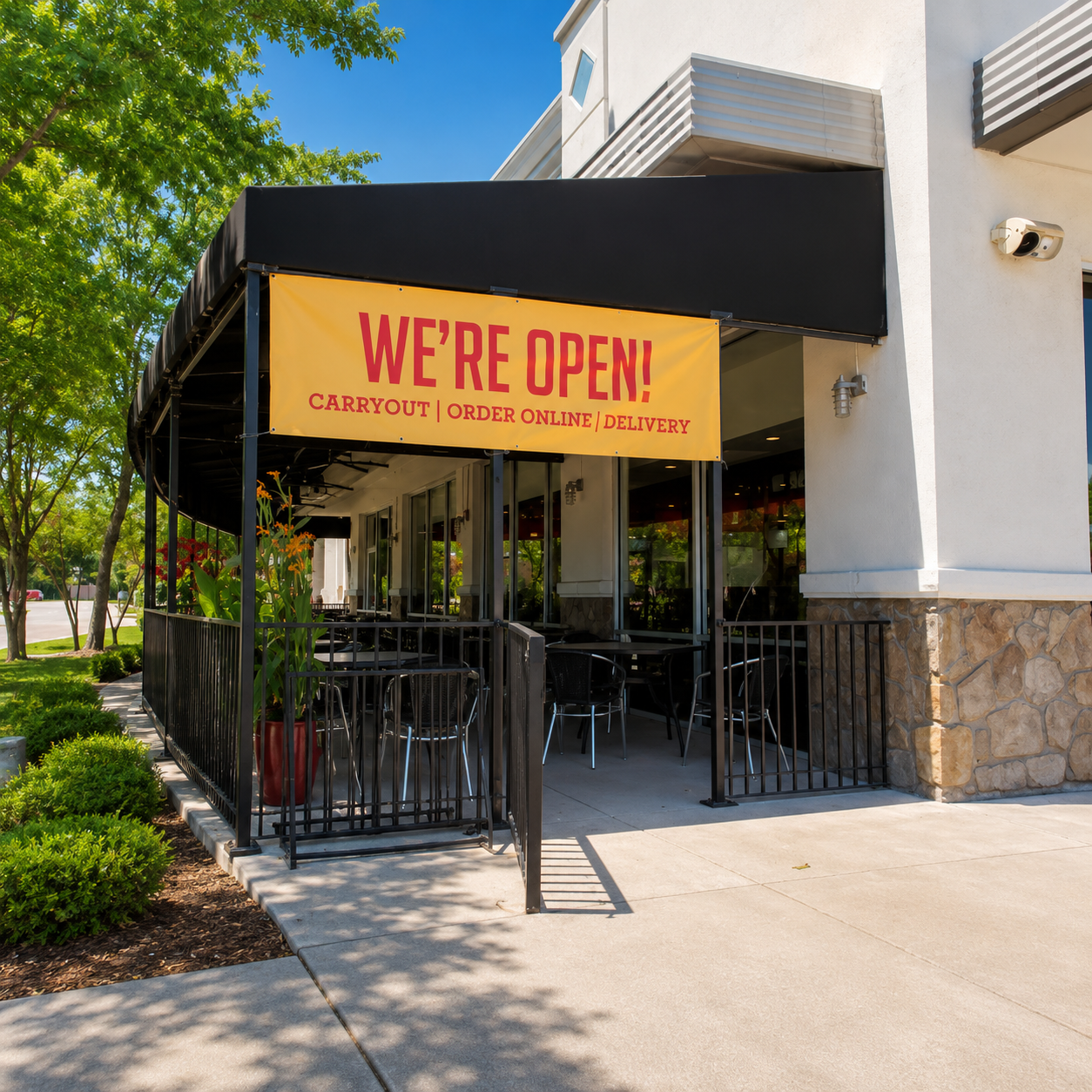 A storefront entrance with a black awning and outdoor seating area under a yellow banner that reads “WE’RE OPEN! Carryout | Order Online | Delivery.” The building has a white exterior with stone base detailing, and the scene is set outdoors with trees, landscaping, and a concrete walkway.