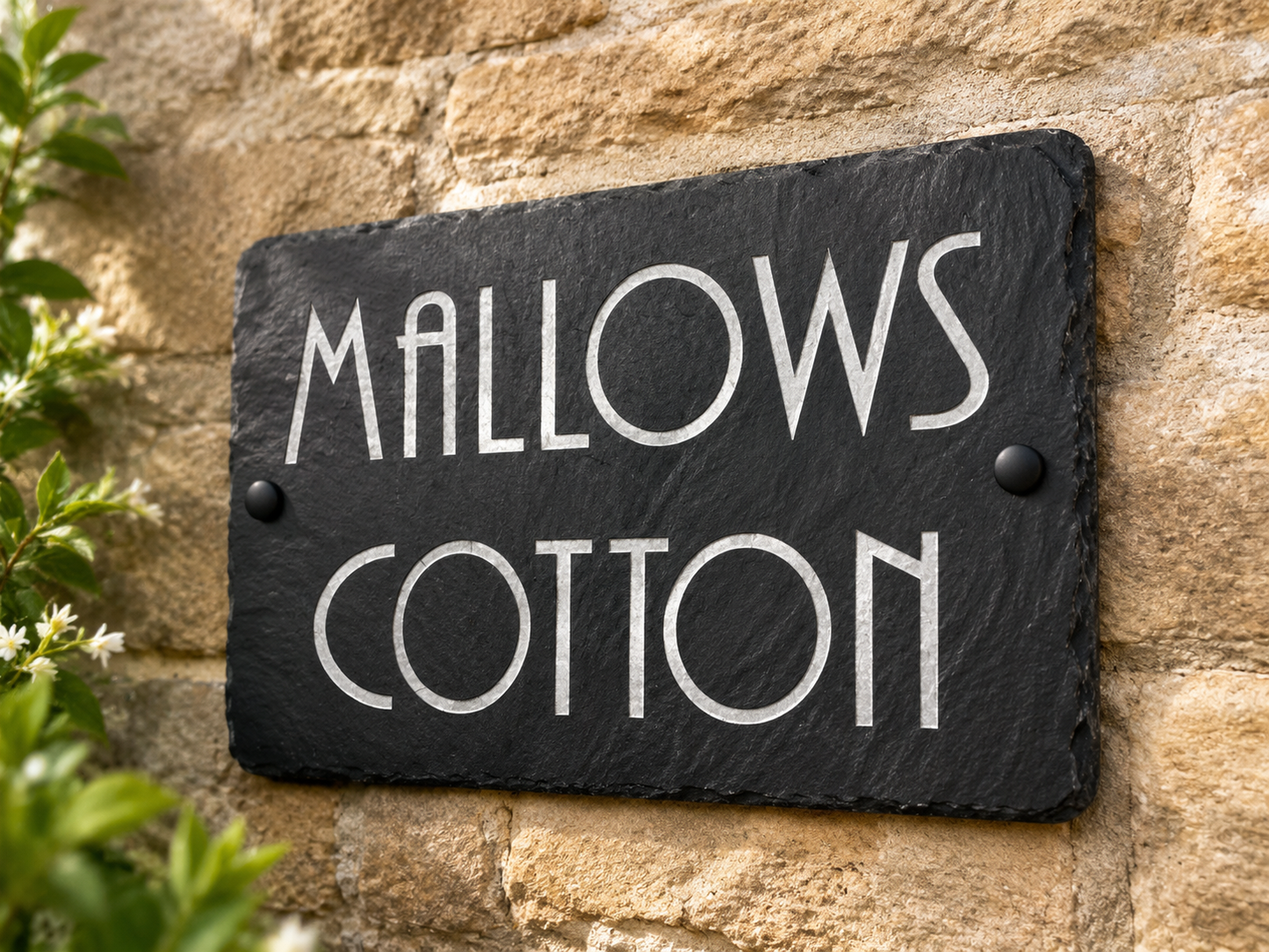 A rectangular black slate sign with white lettering that reads 'MALLOWS COTTON', mounted on a tan stone wall with green leaves visible at the left edge.