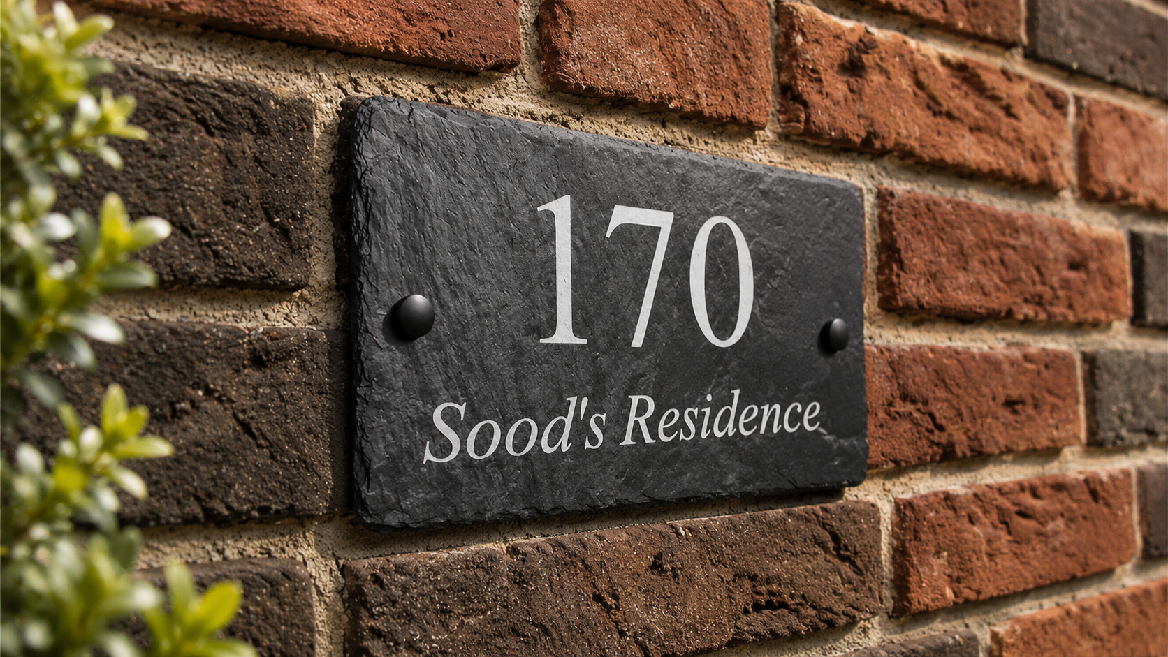A rectangular black slate house number plaque mounted on a red brick wall, with white lettering reading '170 Sood's Residence' and small dark mounting screws on each side.