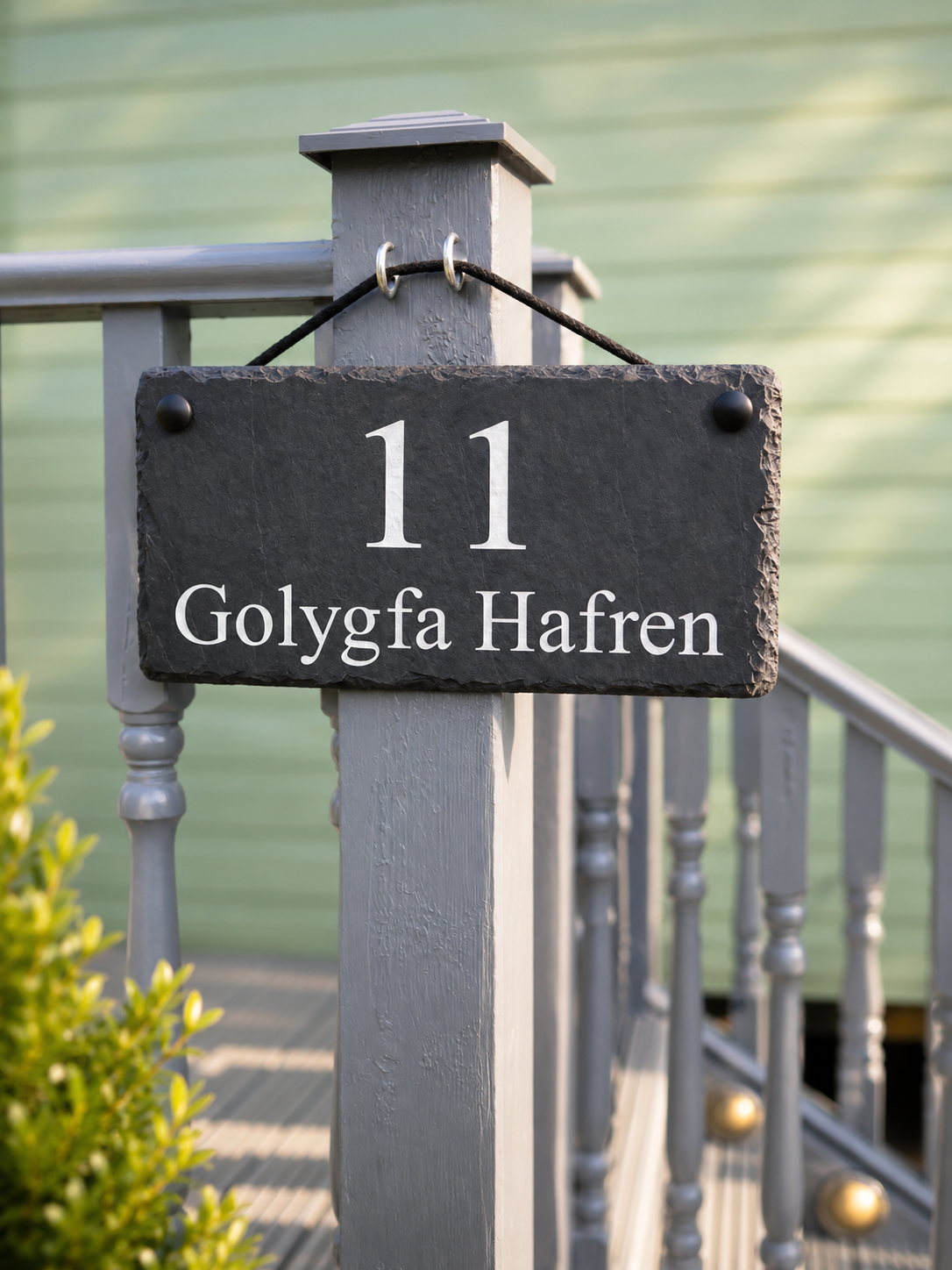 A rectangular dark slate house number sign with white text reading '11 Golygfa Hafren' hanging from a gray wooden post on a porch railing.