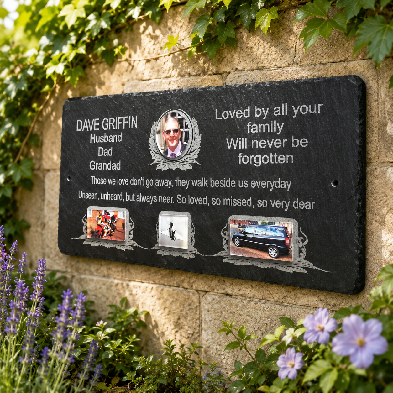Personalized black slate memorial plaque mounted on a stone wall, surrounded by green ivy and purple flowers. The plaque features engraved white text honoring Dave Griffin as husband, dad, and granddad, with a portrait photo, a poem, and three small inset photos.