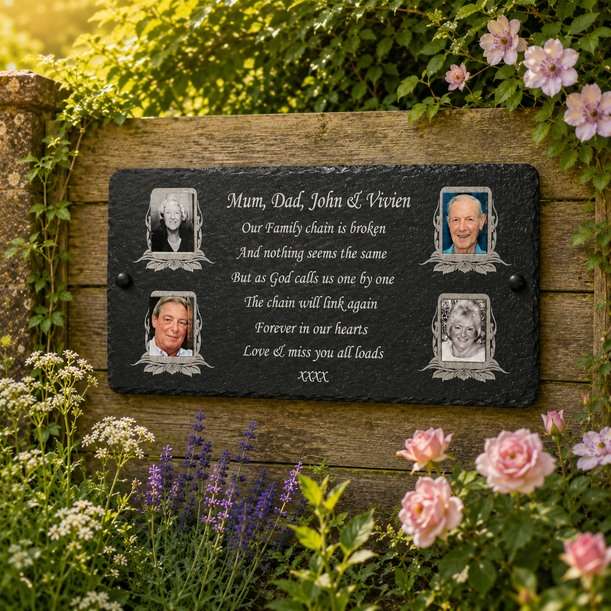 Personalized black memorial garden plaque mounted on a wooden wall in a lush garden setting. The slate plaque features engraved white text with a poem for 'Mum, Dad, John & Vivien' and four portrait photo frames around the text.