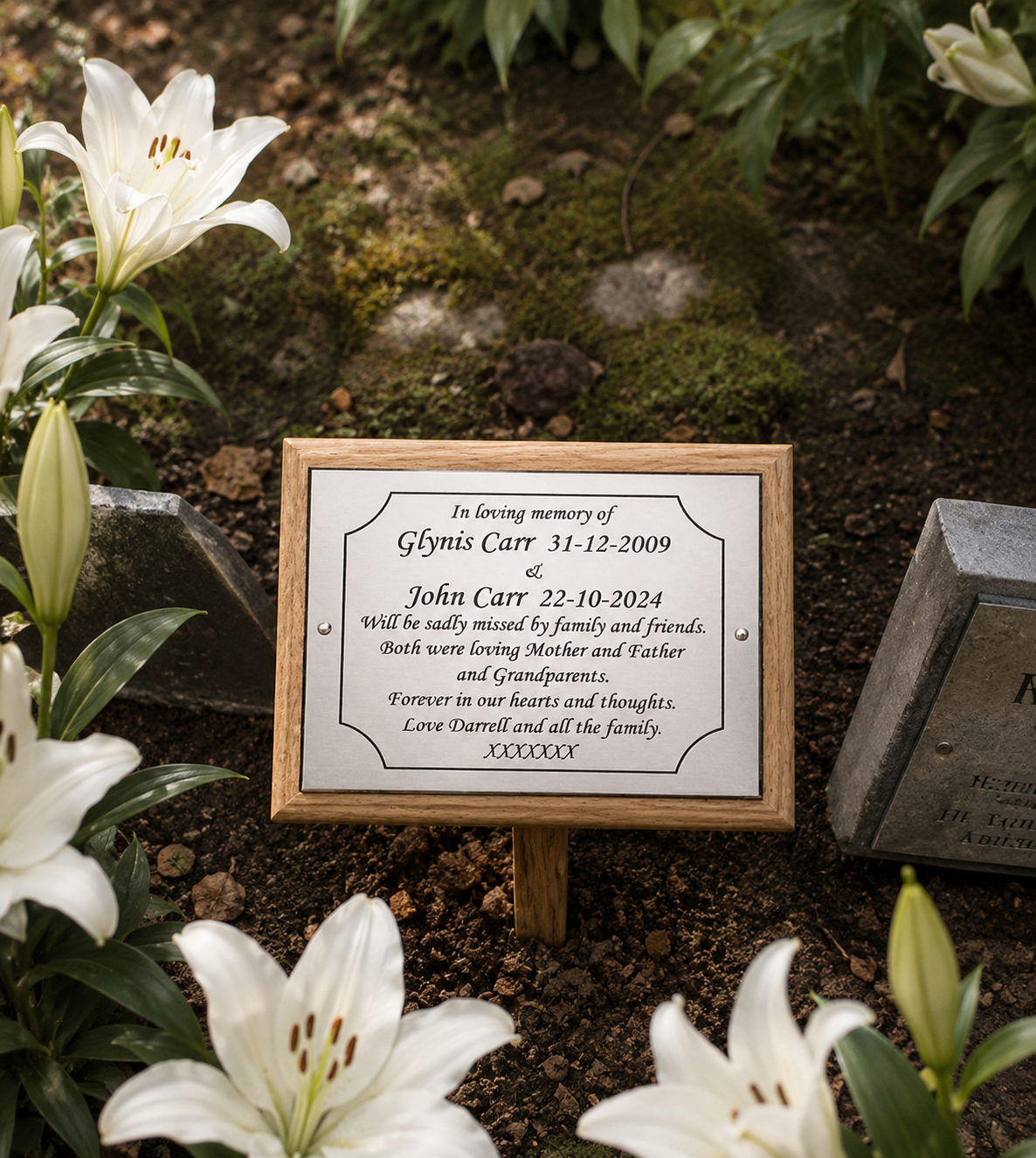 A personalized wooden grave marker plaque with a white printed memorial inscription, displayed outdoors in a garden setting among white lilies and soil.