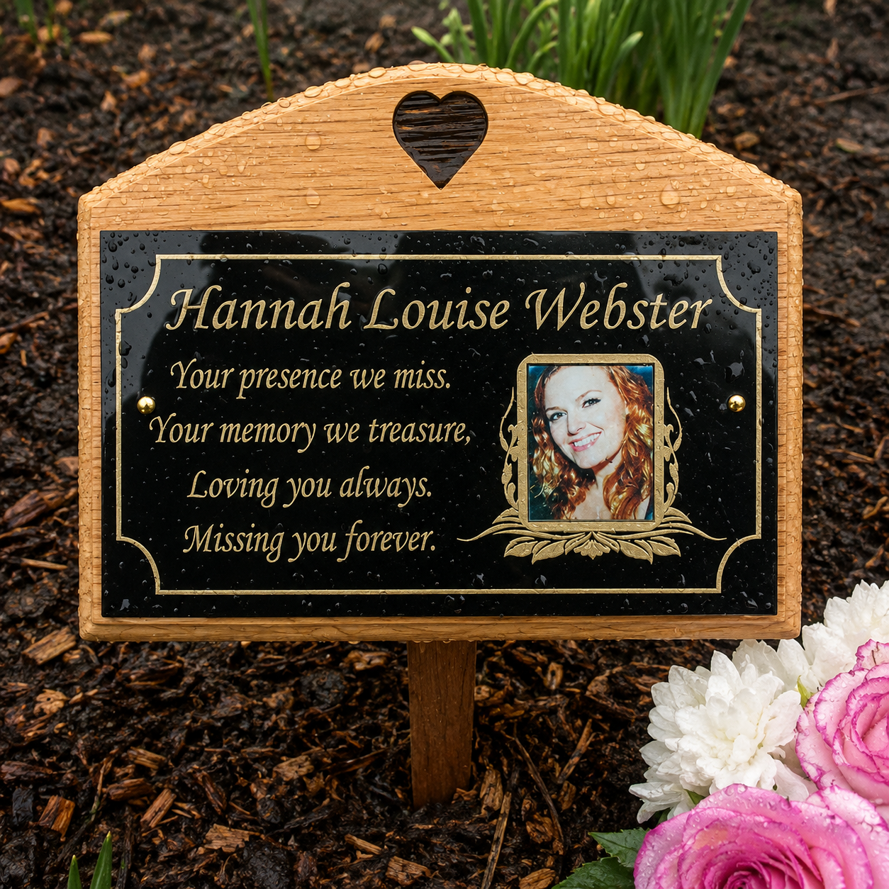 Personalized wooden memorial plaque on a stake, shown outdoors in mulch with flowers nearby. The plaque has a natural wood border, a black center panel with gold lettering, a heart-shaped cutout at the top, and a small portrait photo of a woman.