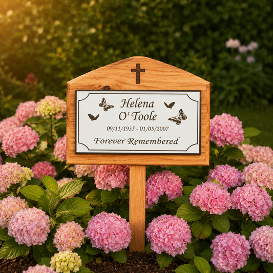 A personalized wooden garden memorial plaque with a cross at the top, surrounded by pink hydrangeas in a garden. The white plaque features black text reading 'Helena O'Toole', the dates '09/11/1935 - 01/05/2007', and 'Forever Remembered', along with small butterfly illustrations.