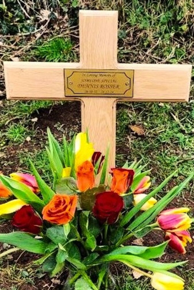 Wooden cross with engraved plaque and colorful flowers on a grassy background