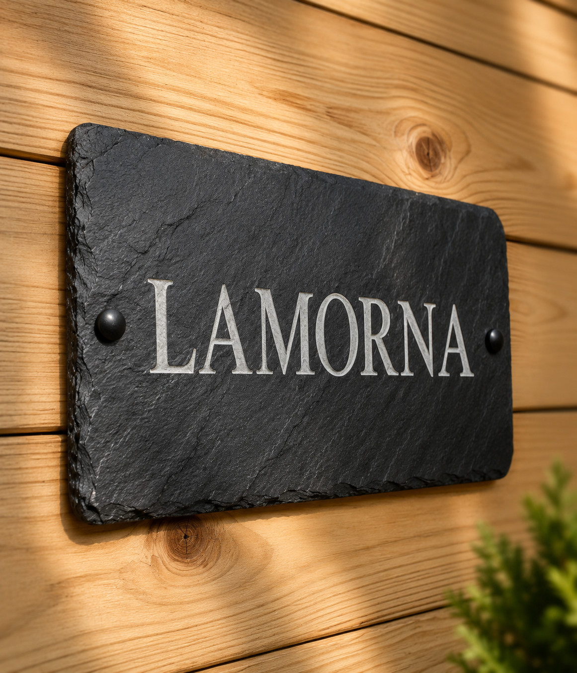 A rectangular black slate house name plaque with the name 'LAMORNA' in white serif lettering, mounted on a light wood plank wall.