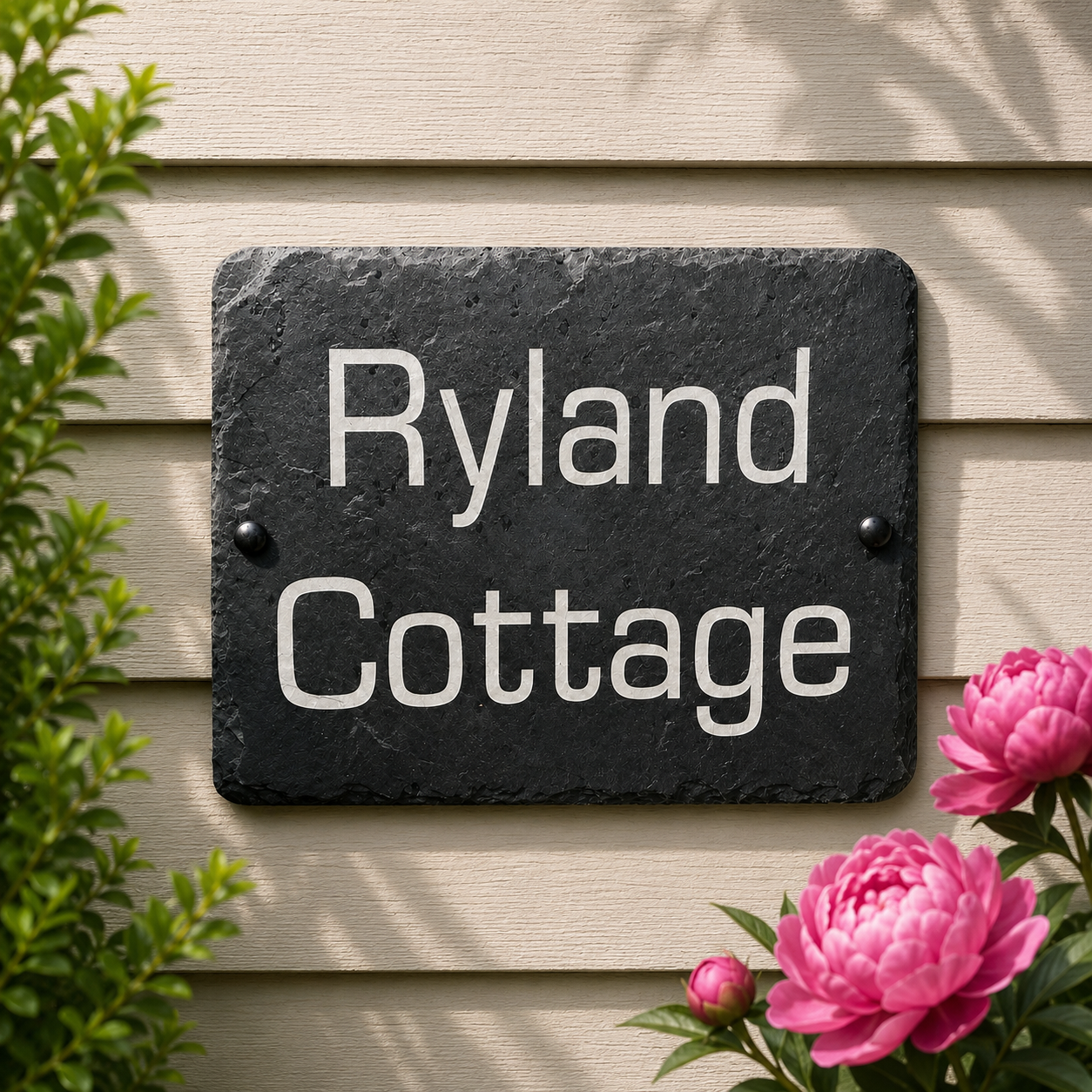 A rectangular black slate house sign with rounded corners and the words 'Ryland Cottage' in white serif lettering, mounted on beige horizontal siding with green leaves and pink flowers around it.