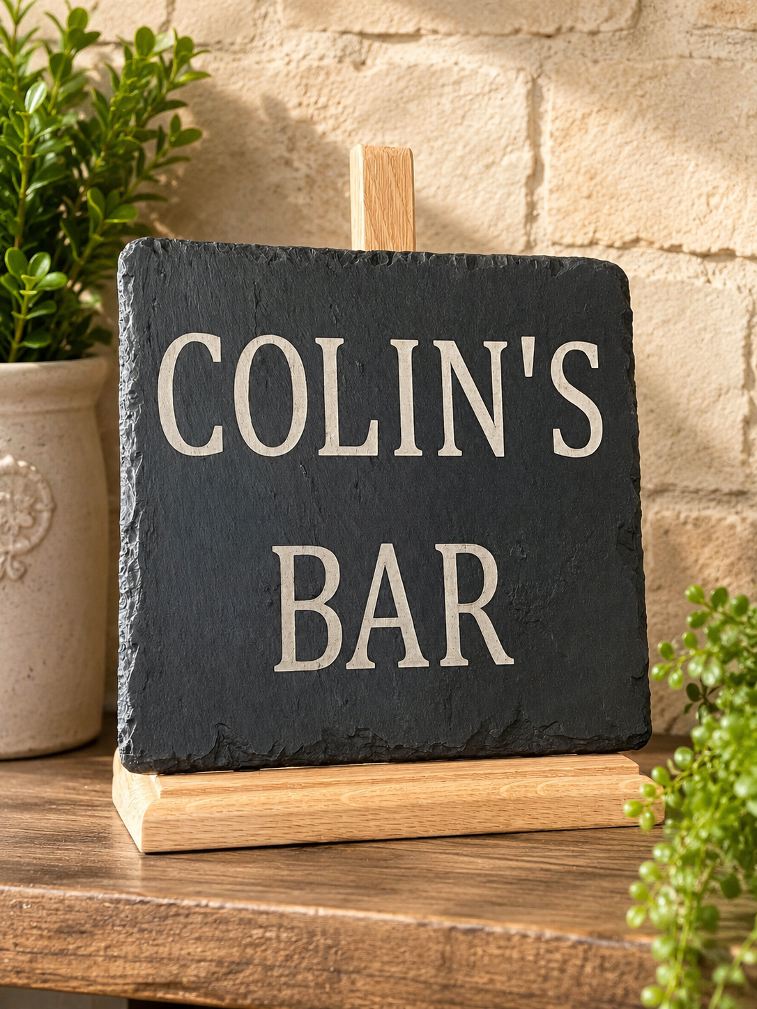A personalized black slate sign reading 'COLIN'S BAR' in raised cream lettering, displayed on a small wooden easel stand on a wooden table with a brick wall and potted plants in the background.