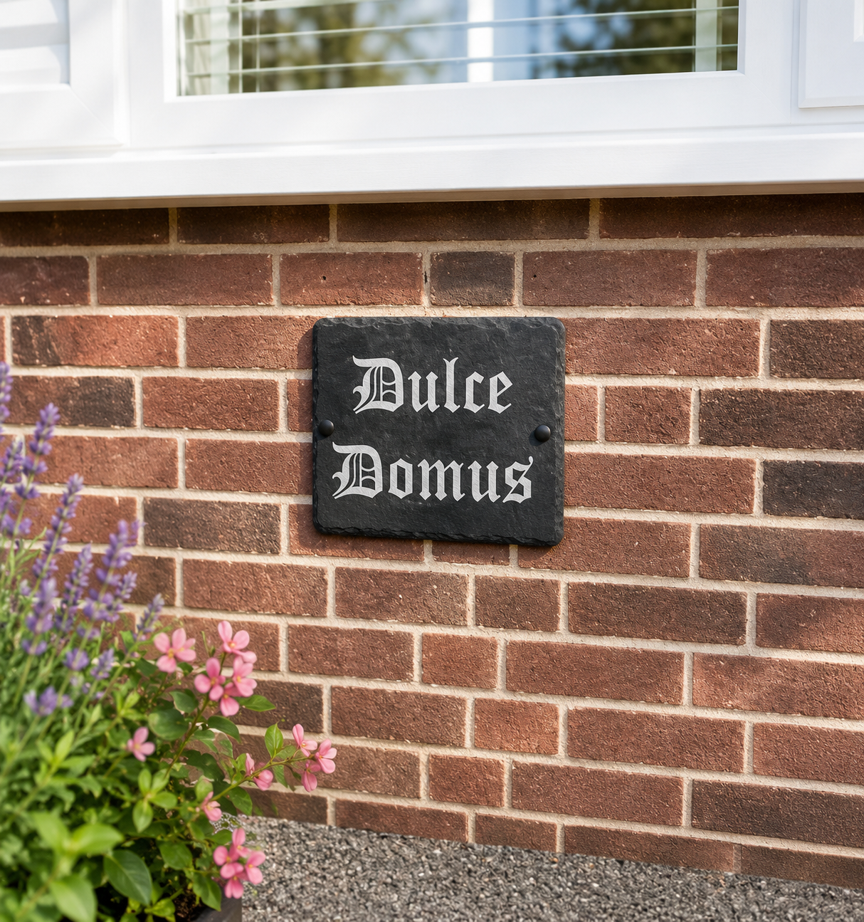 A rectangular black slate house sign with white Gothic lettering reading 'Bulce Domus' mounted on a red brick wall next to purple and pink flowers.