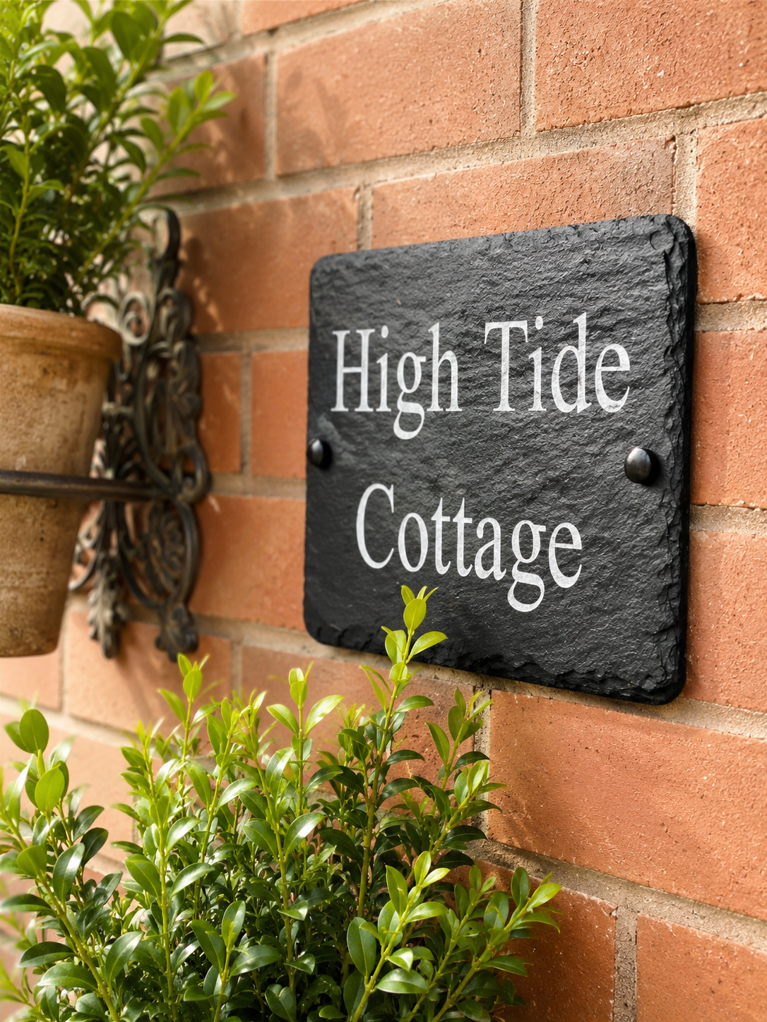 A rectangular black slate house sign with white lettering that reads 'High Tide Cottage,' mounted on a red brick wall with green plants nearby.