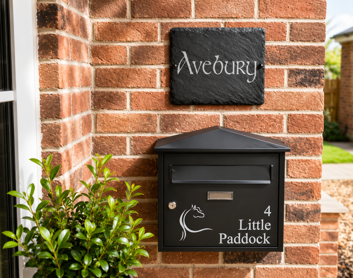 A black metal house mailbox mounted on a red brick wall, with a matching black slate sign above it reading 'Avebury' and the mailbox marked '4 Little Paddock' with a horse silhouette.