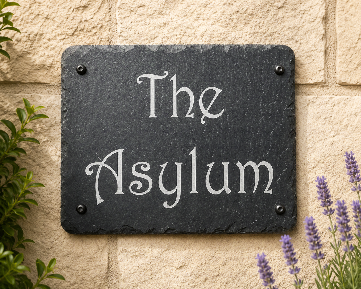 A rectangular black slate sign with the words 'The Asylum' in white script, mounted on a beige stone wall with green leaves and purple flowers at the edges.