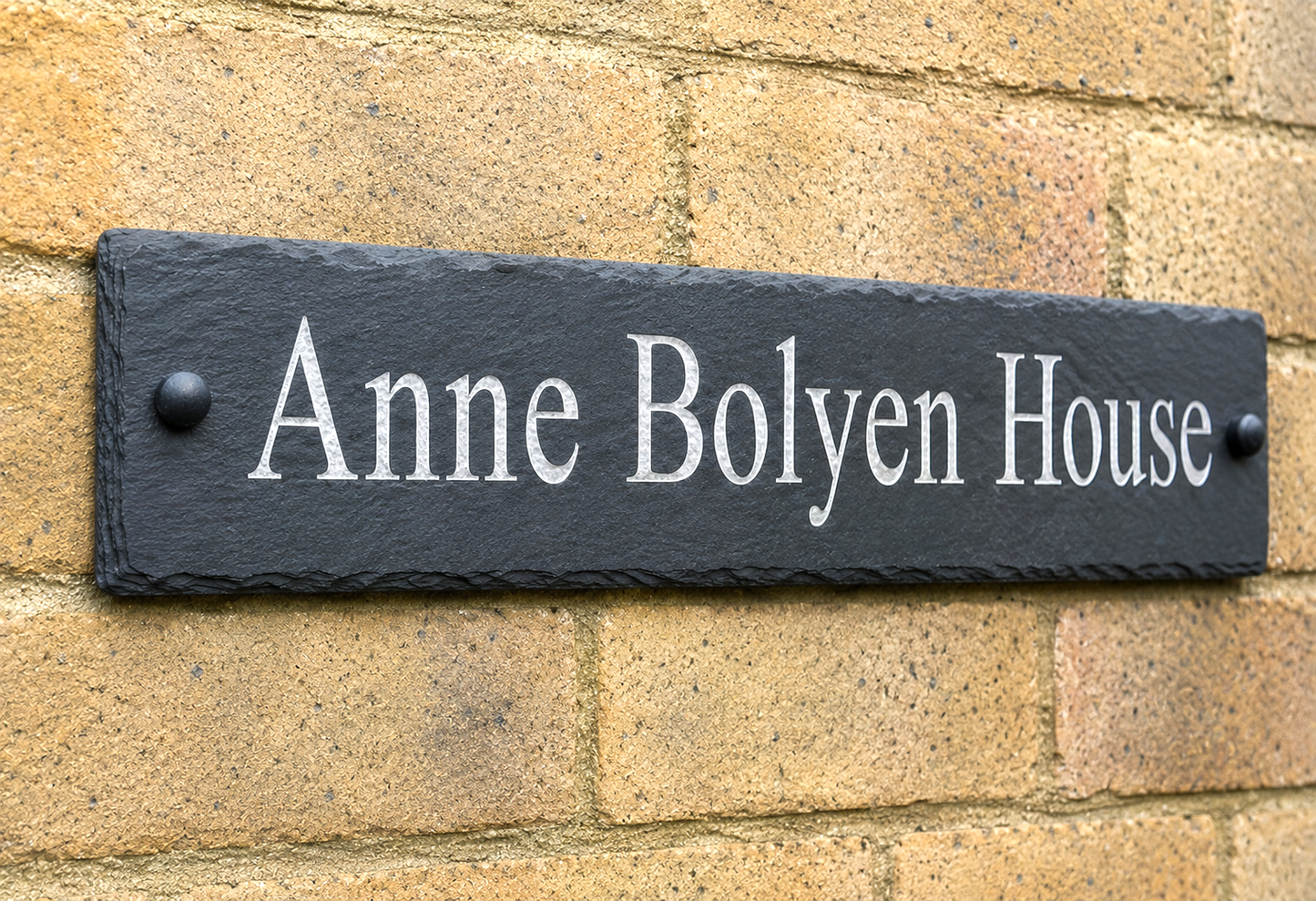 A rectangular black slate house sign with white lettering reading 'Anne Bolyen House', mounted on a tan brick wall.