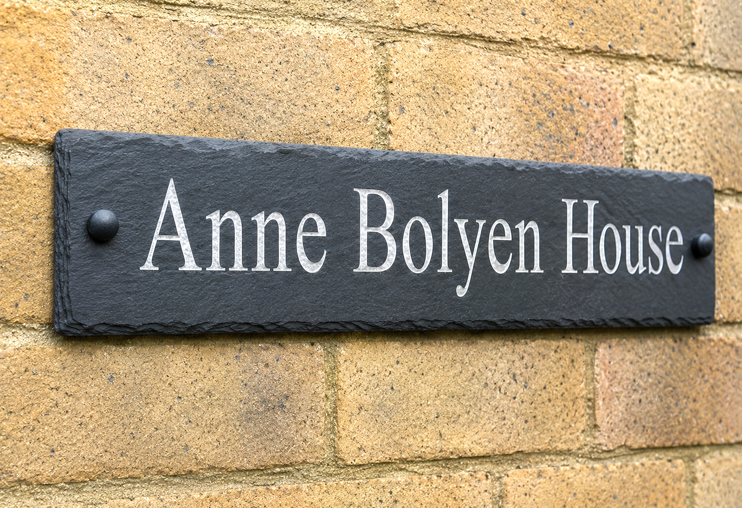 A rectangular black slate house sign with white lettering reading 'Anne Bolyen House', mounted on a tan brick wall.