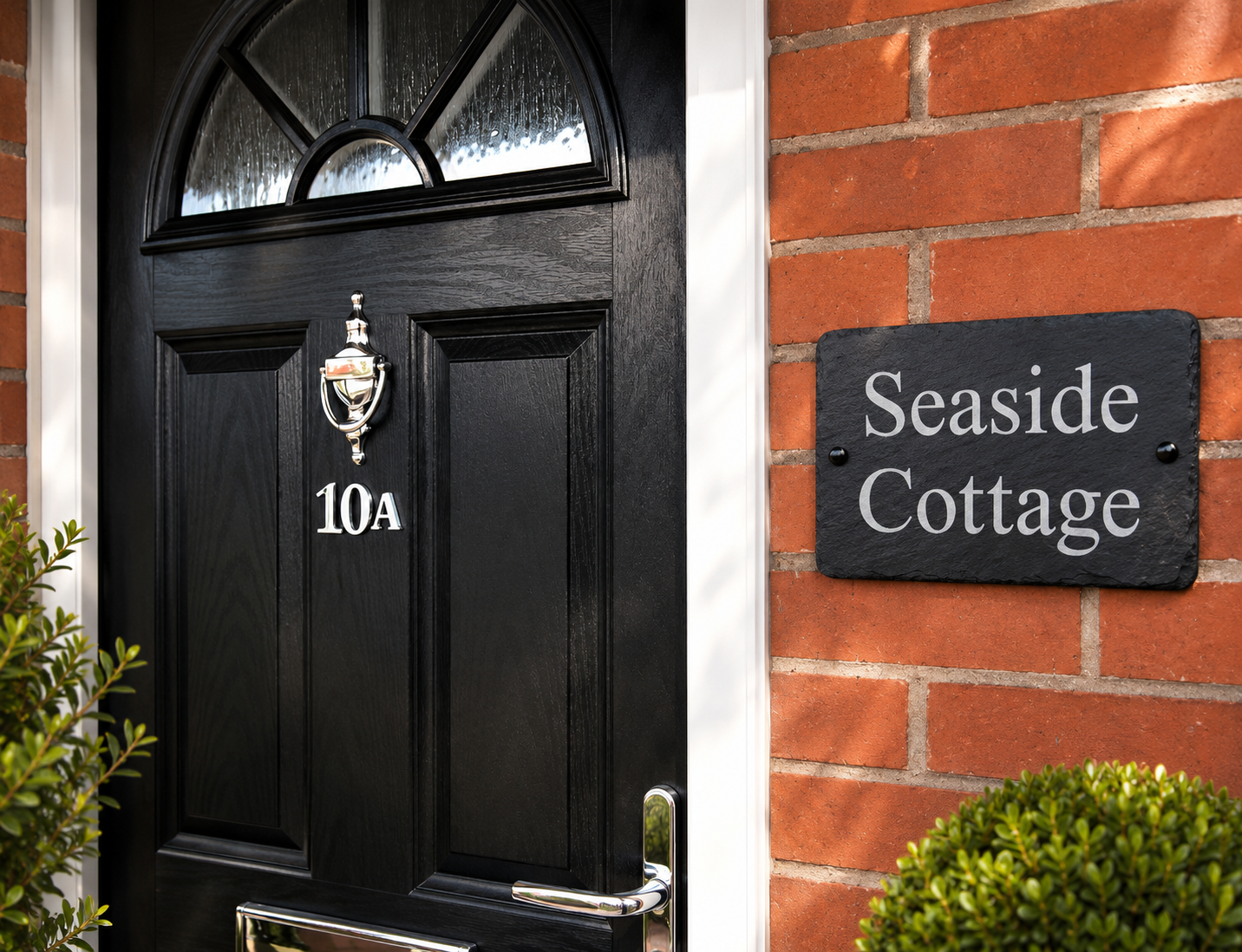 A black front door with a brass knocker and the house number 10A, set against a red brick wall with a black slate sign reading 'Seaside Cottage'.