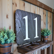 Slate number '11' sign on a wooden shelf with plants against a wooden wall.