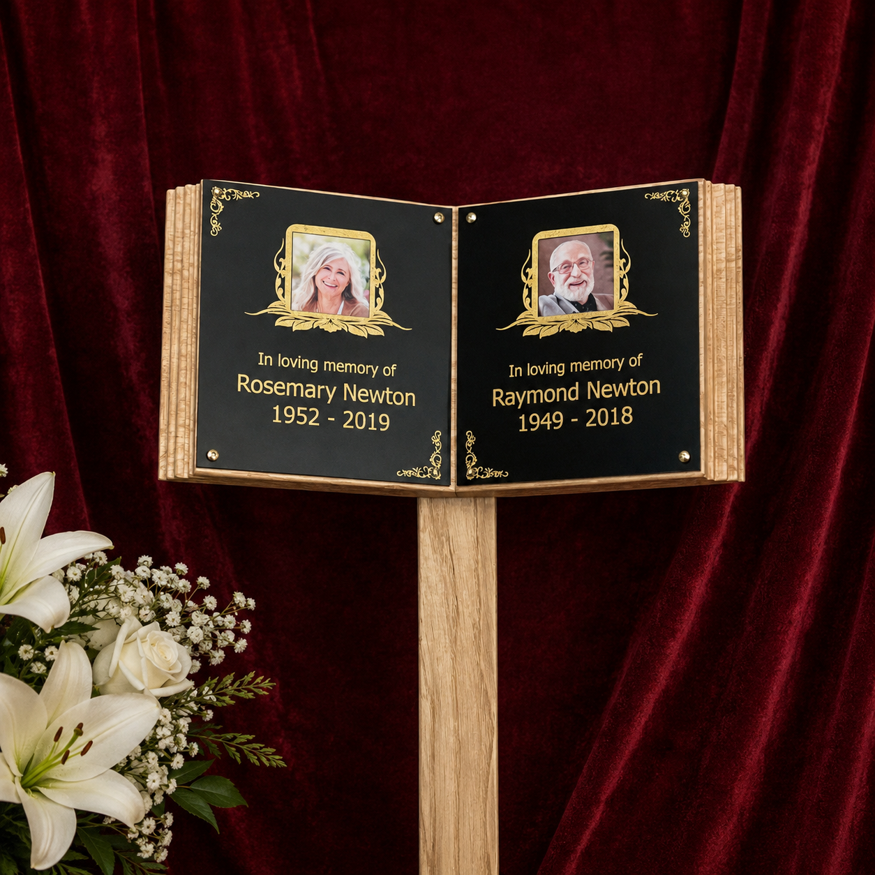 A wooden memorial book grave marker on a wooden stand, featuring black panels with gold lettering and portrait frames, set against a dark red velvet curtain with white flowers in the foreground.