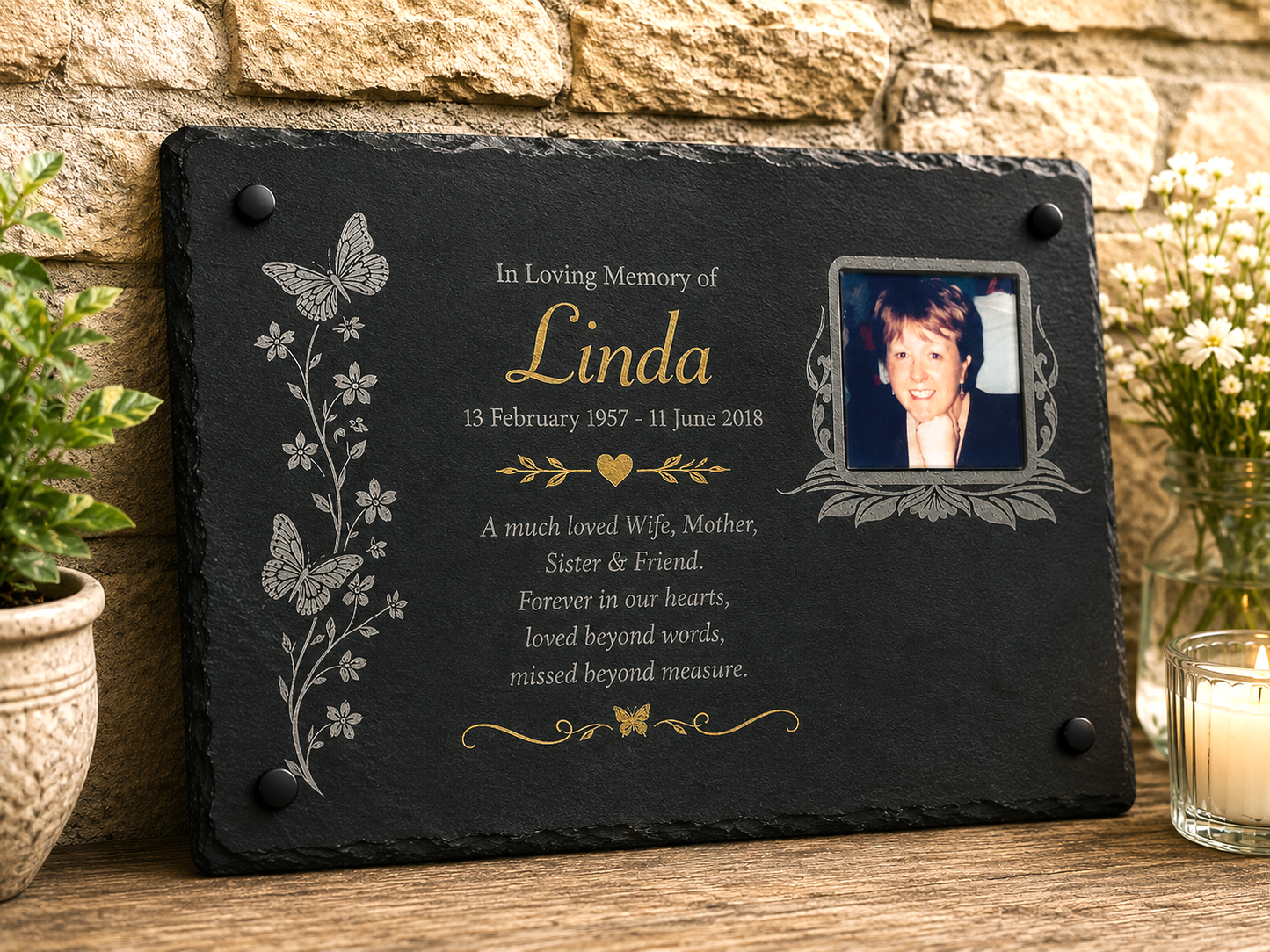 Personalized black slate memorial plaque with engraved gold and silver text, floral and butterfly motifs, a photo frame, and a candle on a wooden surface against a stone wall.