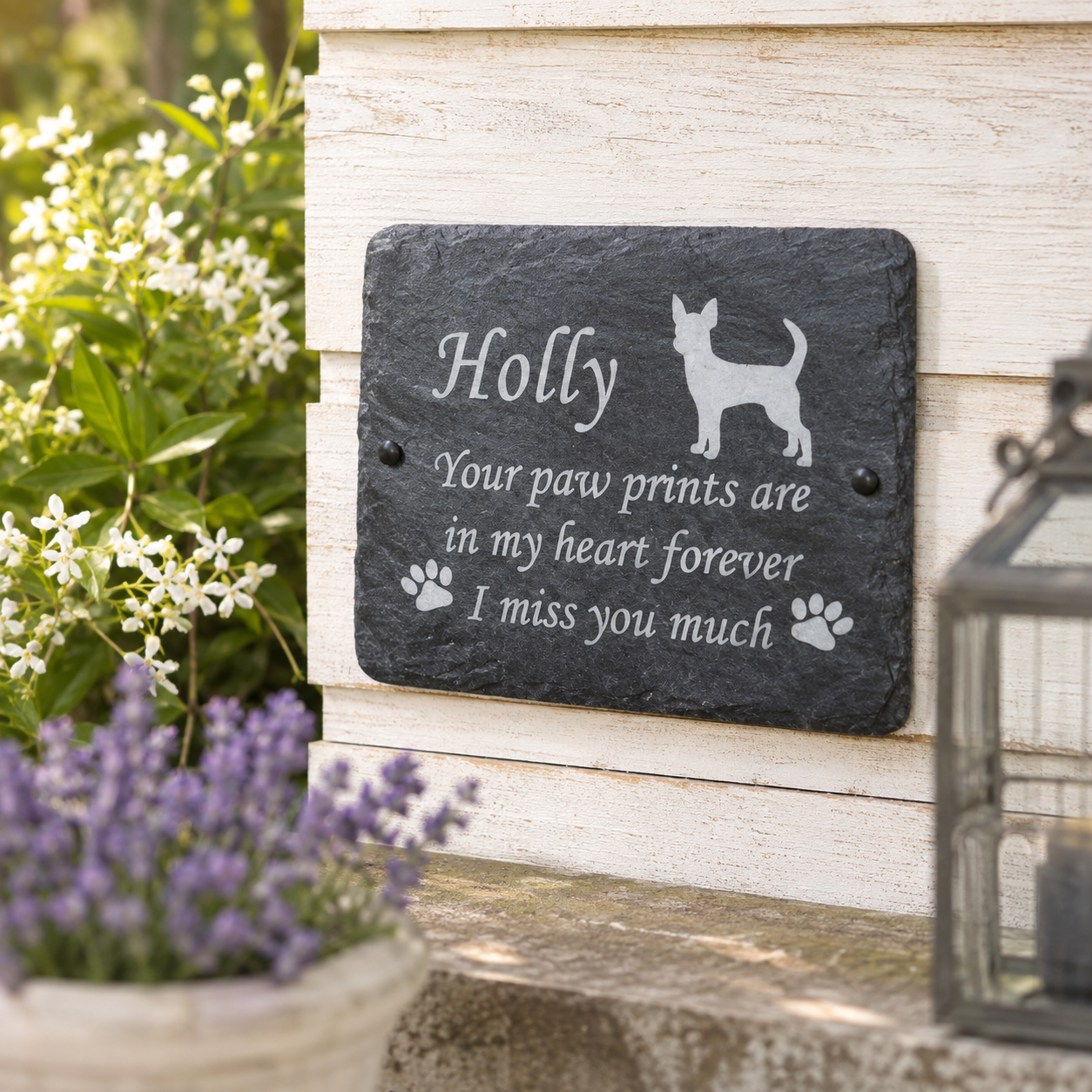 Personalized dark slate plaque with engraved white text reading 'Holly Your paw prints are in my heart forever I miss you much' and a dog silhouette, mounted on a light wood wall with garden plants and a lantern in the background.