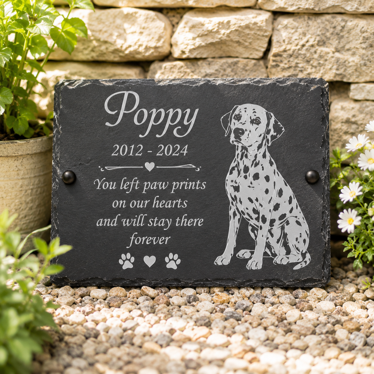 Personalized black slate pet memorial plaque engraved with the name 'Poppy', dates '2012 - 2024', a remembrance quote, paw prints, and a Dalmatian illustration, displayed outdoors on gravel against a stone wall with plants.