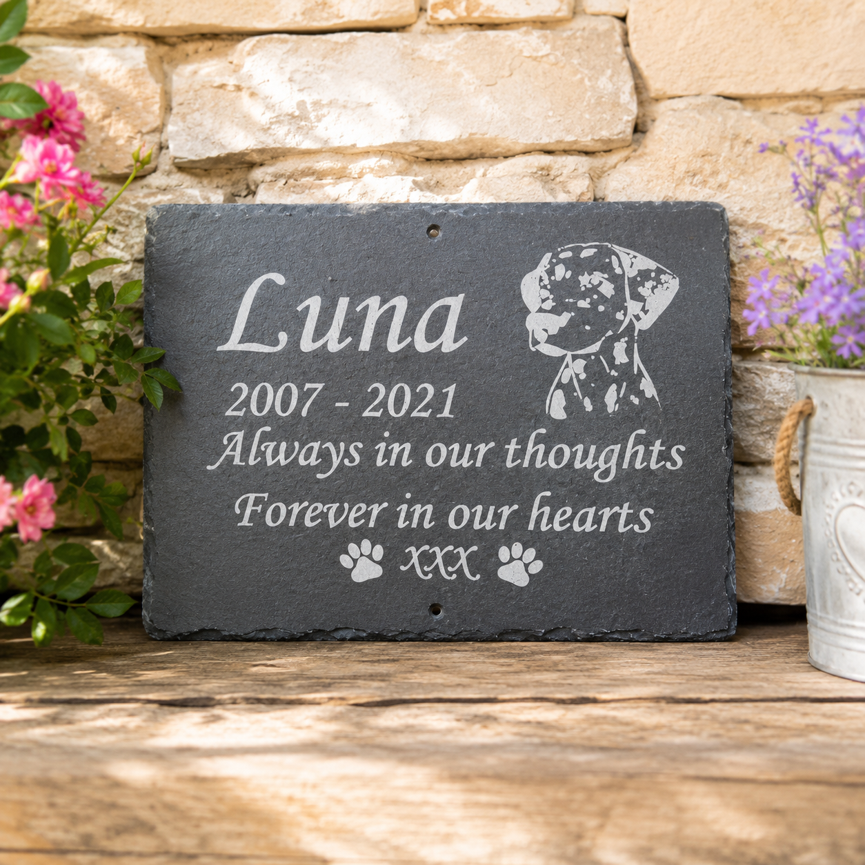 Personalized dark gray slate memorial plaque with engraved white text reading 'Luna 2007 - 2021 Always in our thoughts Forever in our hearts' plus paw prints, shown leaning on a wooden surface against a stone wall with flowers.