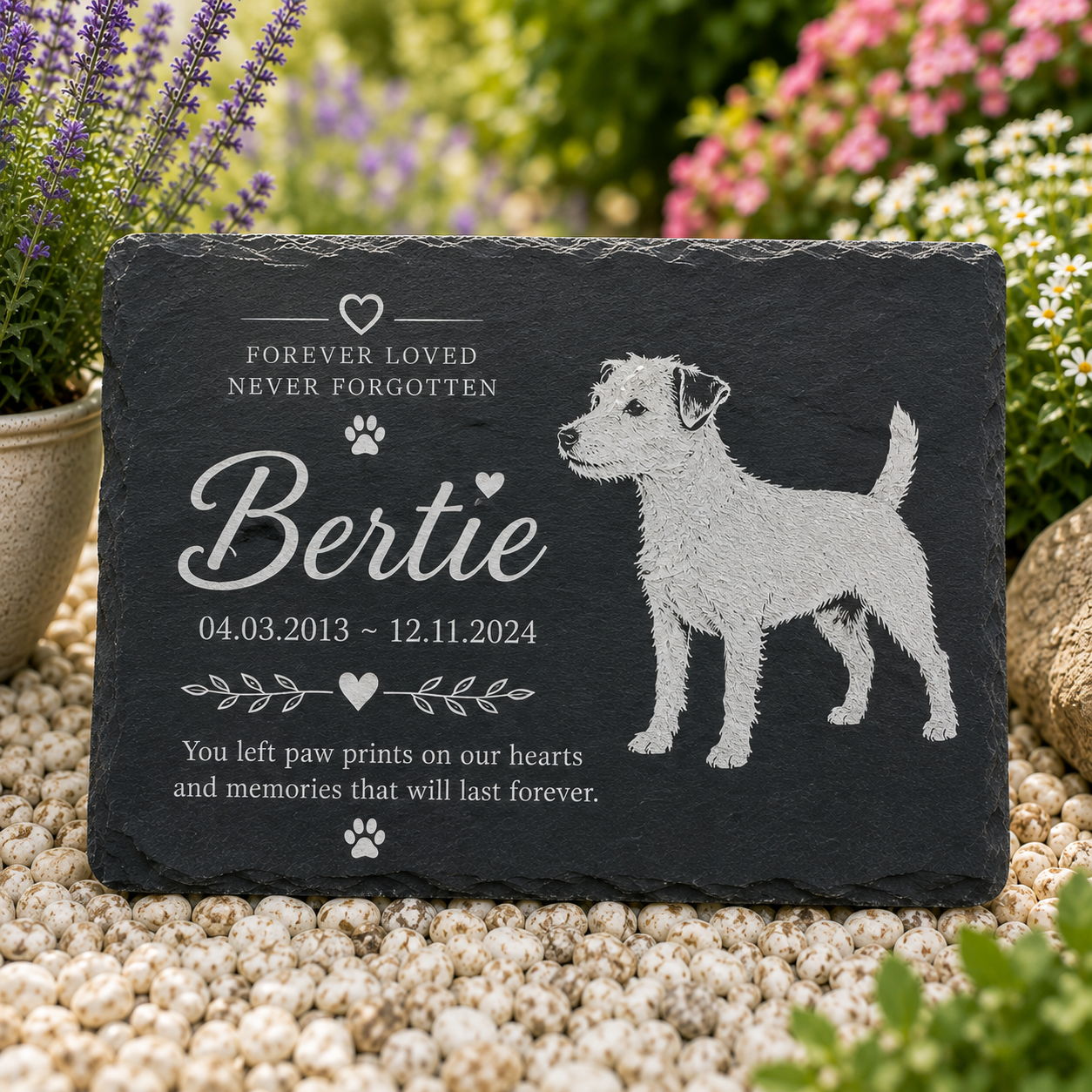 Personalized black slate pet memorial plaque engraved with the name 'Bertie', dates, paw prints, and a dog silhouette, displayed outdoors on pebbles with flowers in the background.