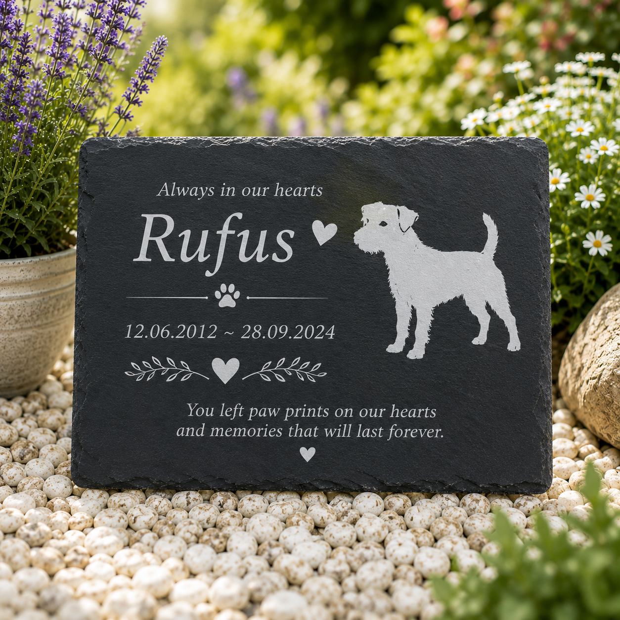 Personalized dark gray slate pet memorial garden stone engraved with the name 'Rufus', dates, paw prints, and a dog silhouette, displayed on white pebbles with blurred garden plants in the background.