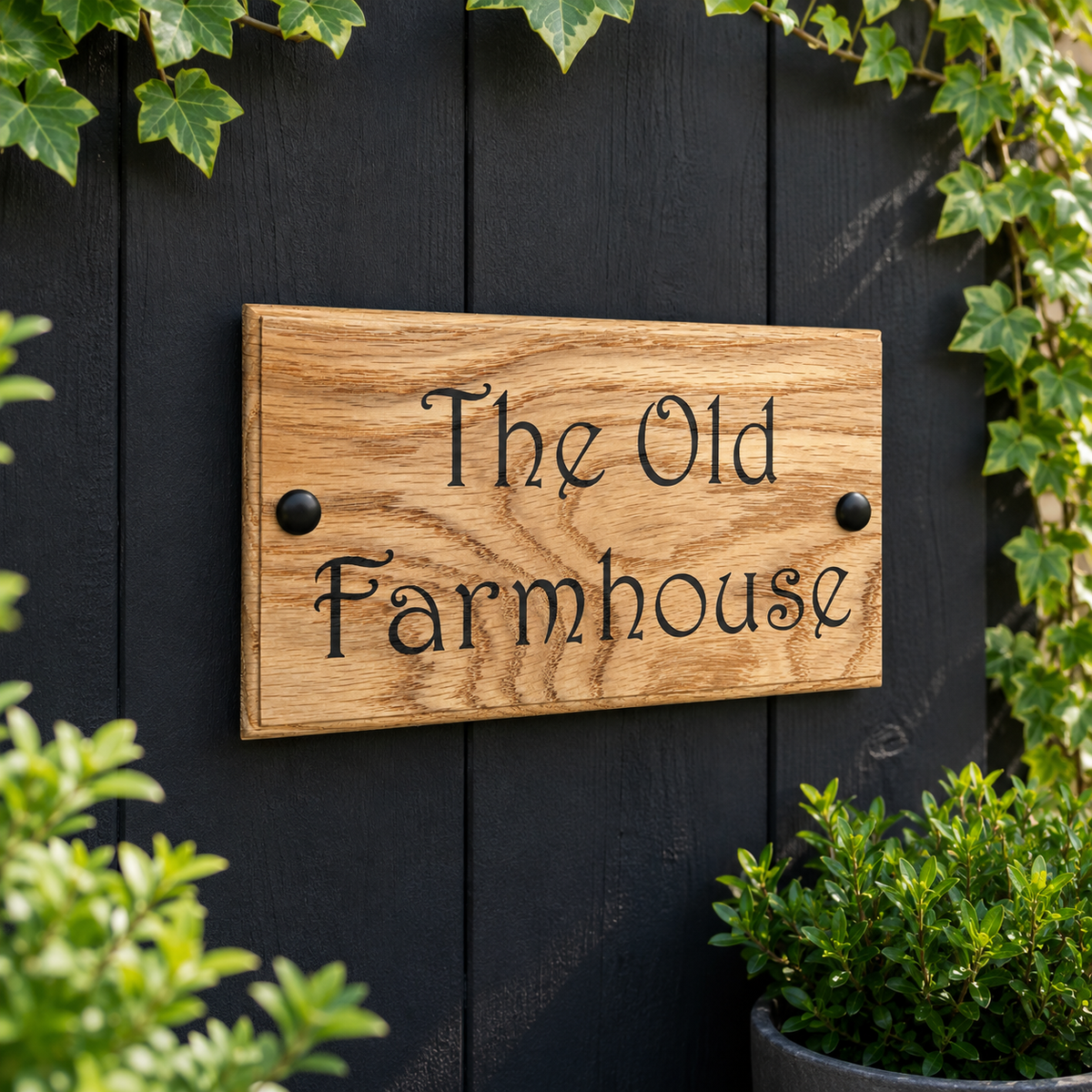 A rectangular wooden sign with the words 'The Old Farmhouse' in black script, mounted on a dark blackboard wall with green ivy and plants around it.