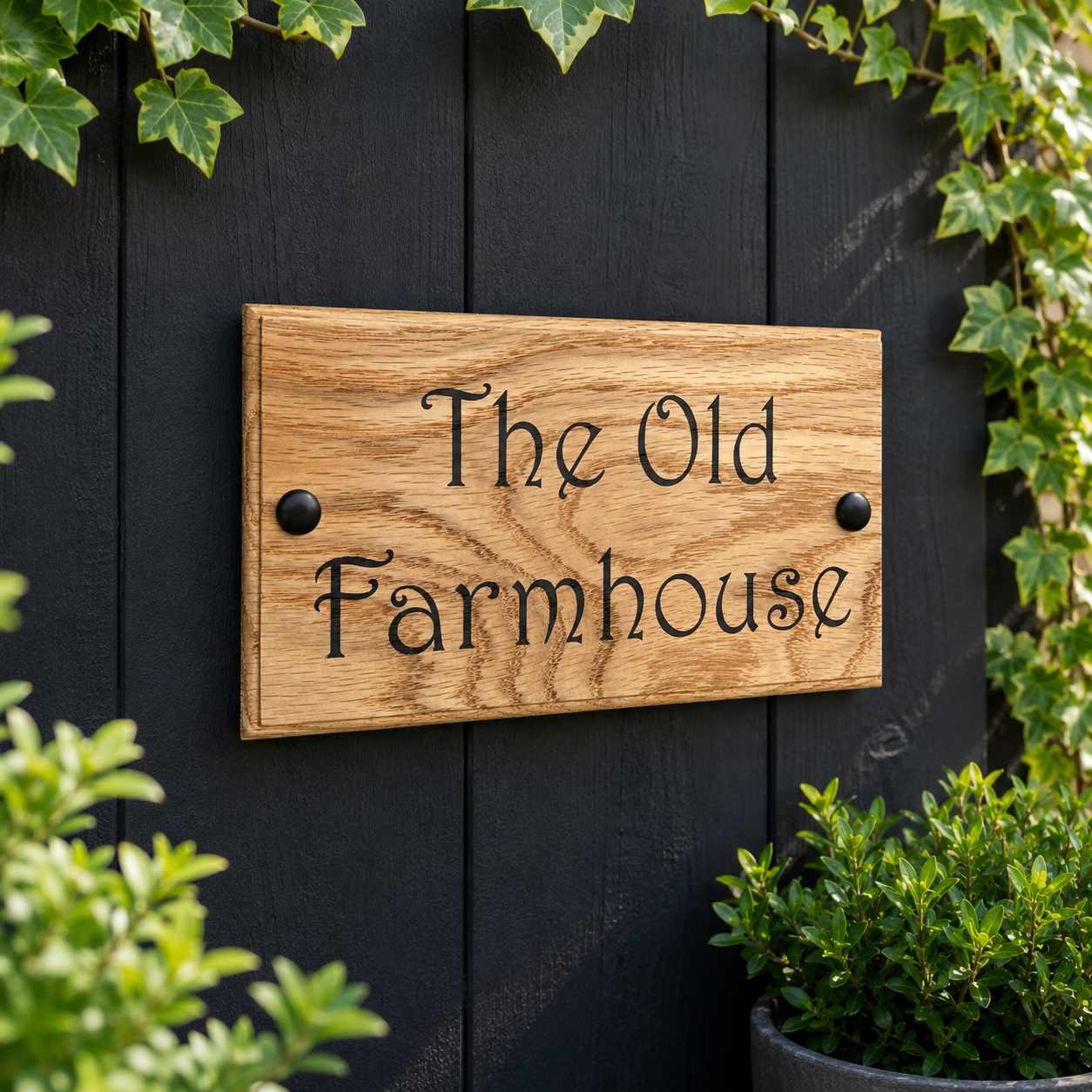A rectangular wooden sign with the words 'The Old Farmhouse' in black script, mounted on a dark blackboard wall with green ivy and plants around it.