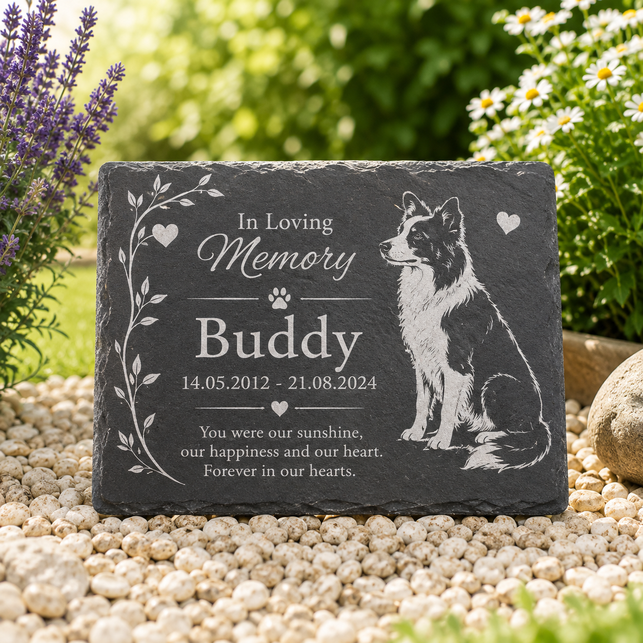 Personalized dark gray slate pet memorial stone engraved with 'In Loving Memory Buddy' and a dog illustration, displayed outdoors on white pebbles with blurred garden plants in the background.