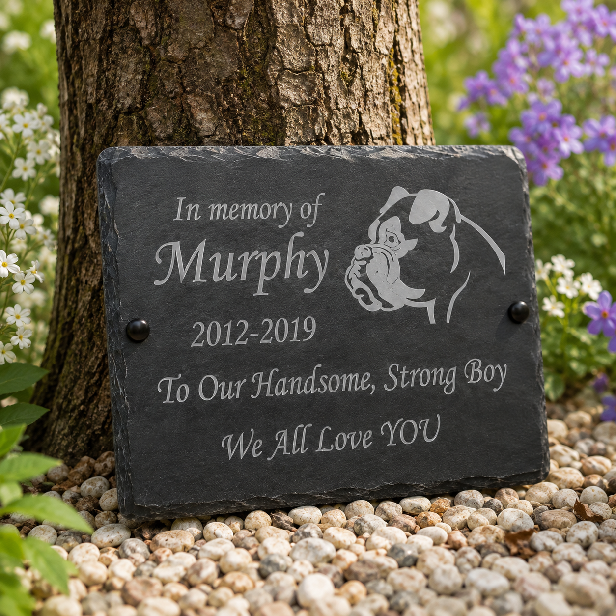 Personalized dark gray slate memorial plaque with engraved text reading 'In memory of Murphy 2012-2019 To Our Handsome, Strong Boy We All Love YOU' and a bulldog silhouette, displayed outdoors against a tree trunk on pebbles with flowers in the background.
