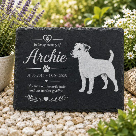 Personalized black slate pet memorial garden stone engraved with a dog silhouette, paw print, and the text 'In loving memory of Archie' with dates and remembrance message, displayed on white pebbles with plants in the background.